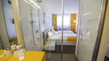 Habitación de hotel con cabina de ducha de vidrio, tocador amarillo, cama con ropa blanca y vistas de la habitación contigua.