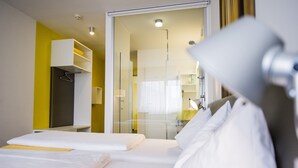 A hotel room with a bed, a bedside table, a lamp, and a glass door leading to a bathroom. - STAY.inn Comfort Art Hotel Schwaz (Schwaz)