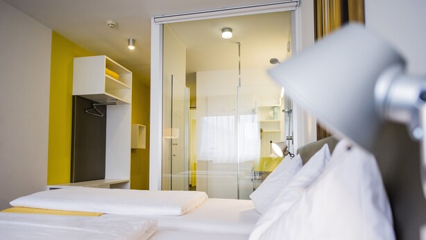 A hotel room with a bed, a bedside table, a lamp, and a glass door leading to a bathroom. - STAY.inn Comfort Art Hotel Schwaz (Schwaz)