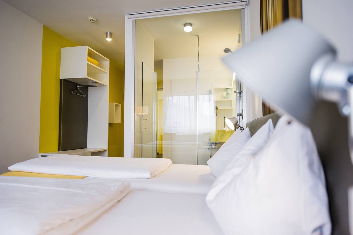 A hotel room with a bed, a bedside table, a lamp, and a glass door leading to a bathroom.