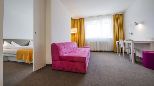A hotel room with a pink sofa, a white bed, a desk, and a chair. - STAY.inn Comfort Art Hotel Schwaz (Schwaz)