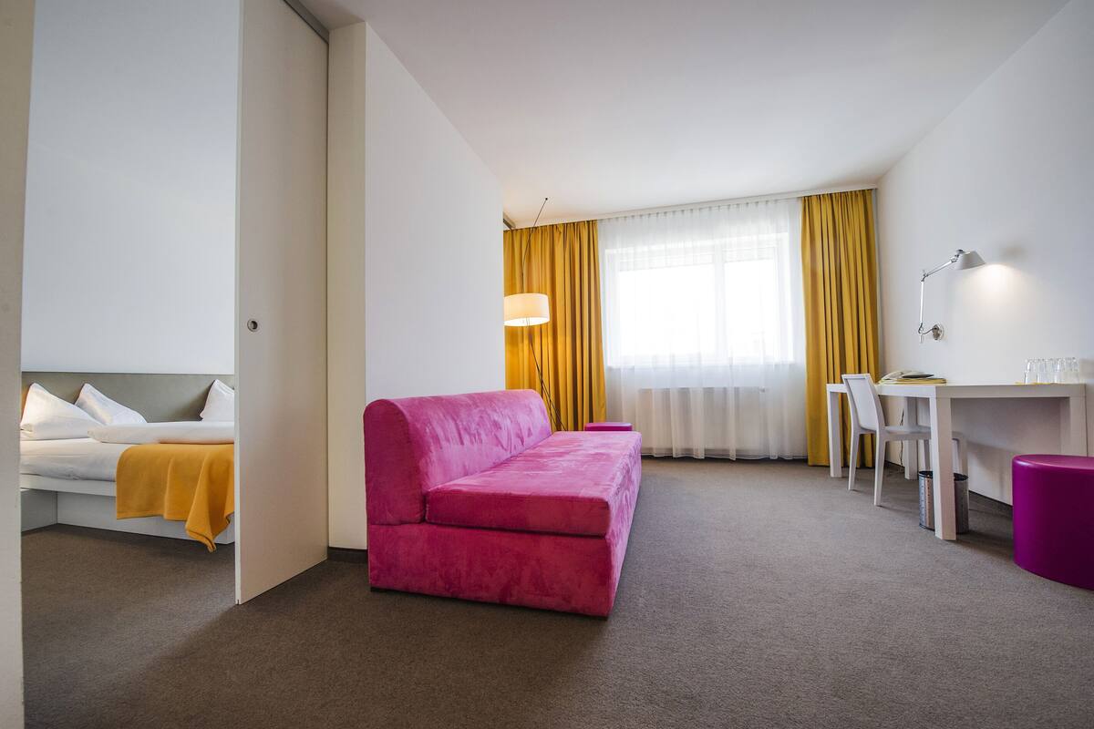 A hotel room with a pink sofa, a white bed, a desk, and a chair.