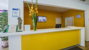 A reception desk with a yellow accent wall, a clock, and a television screen. - STAY.inn Comfort Art Hotel Schwaz (Schwaz)