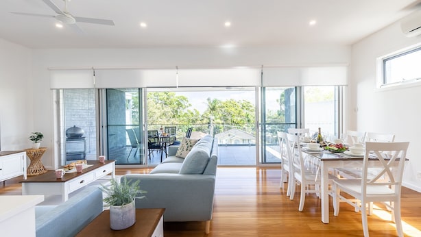 Un amplio salón con sofá gris, mesa de comedor con sillas blancas y un balcón con vistas a árboles y una casa.