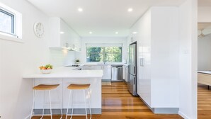 Una cocina moderna con armarios blancos, una isla central con taburetes de bar, electrodomésticos de acero inoxidable y un ventanal que proporciona luz natural.