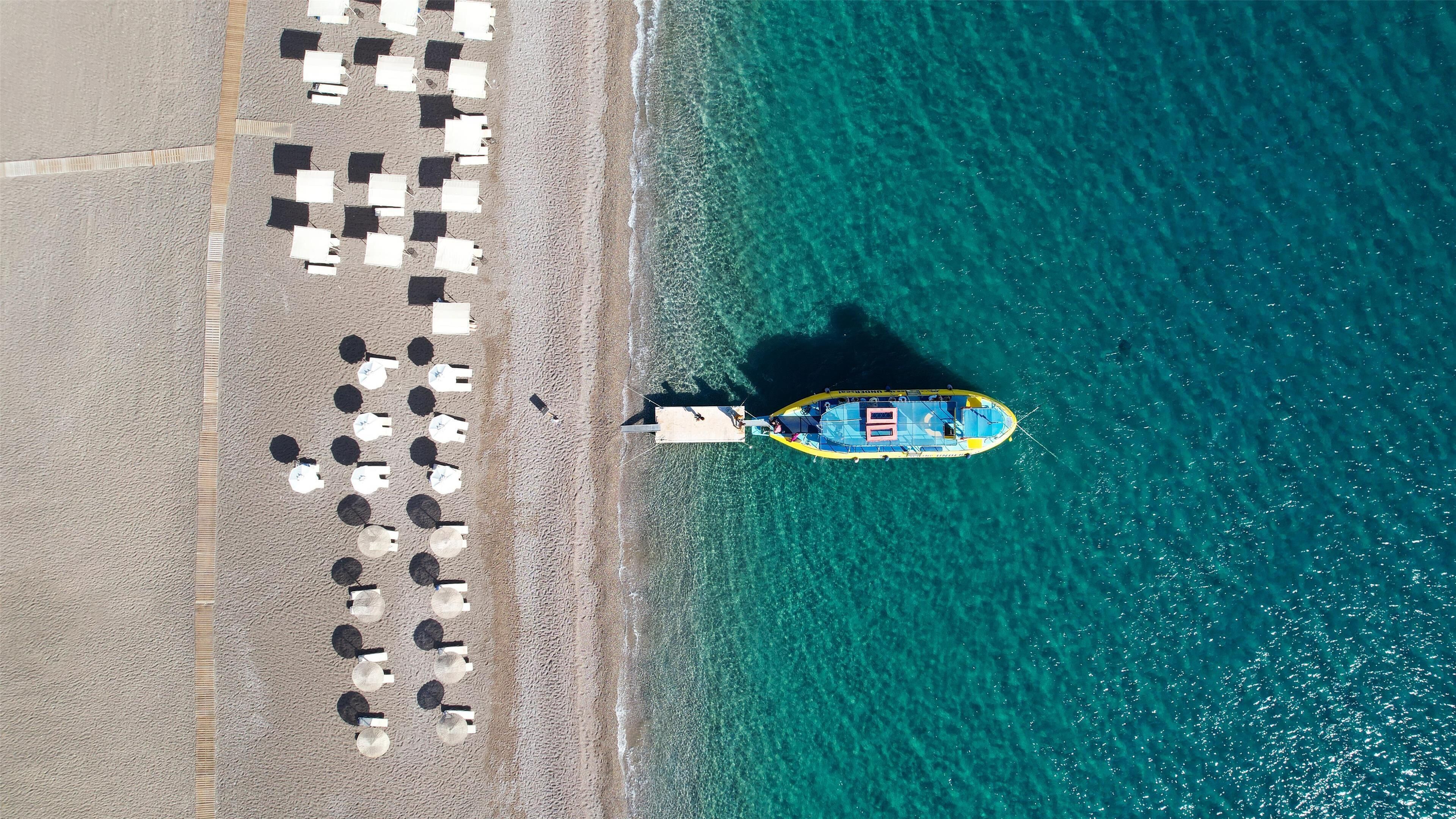An aerial view of a beach with a boat docked near the shore, surrounded by rows of beach umbrellas and chairs.