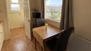 Een kleine kamer met een houten tafel, twee stoelen, een raam met gordijnen, een radiator en uitzicht op een stadsgezicht.