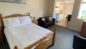 Een slaapkamer met een houten bed, een zwarte bank, een schilderij aan de muur en uitzicht op de keuken en het balkon.