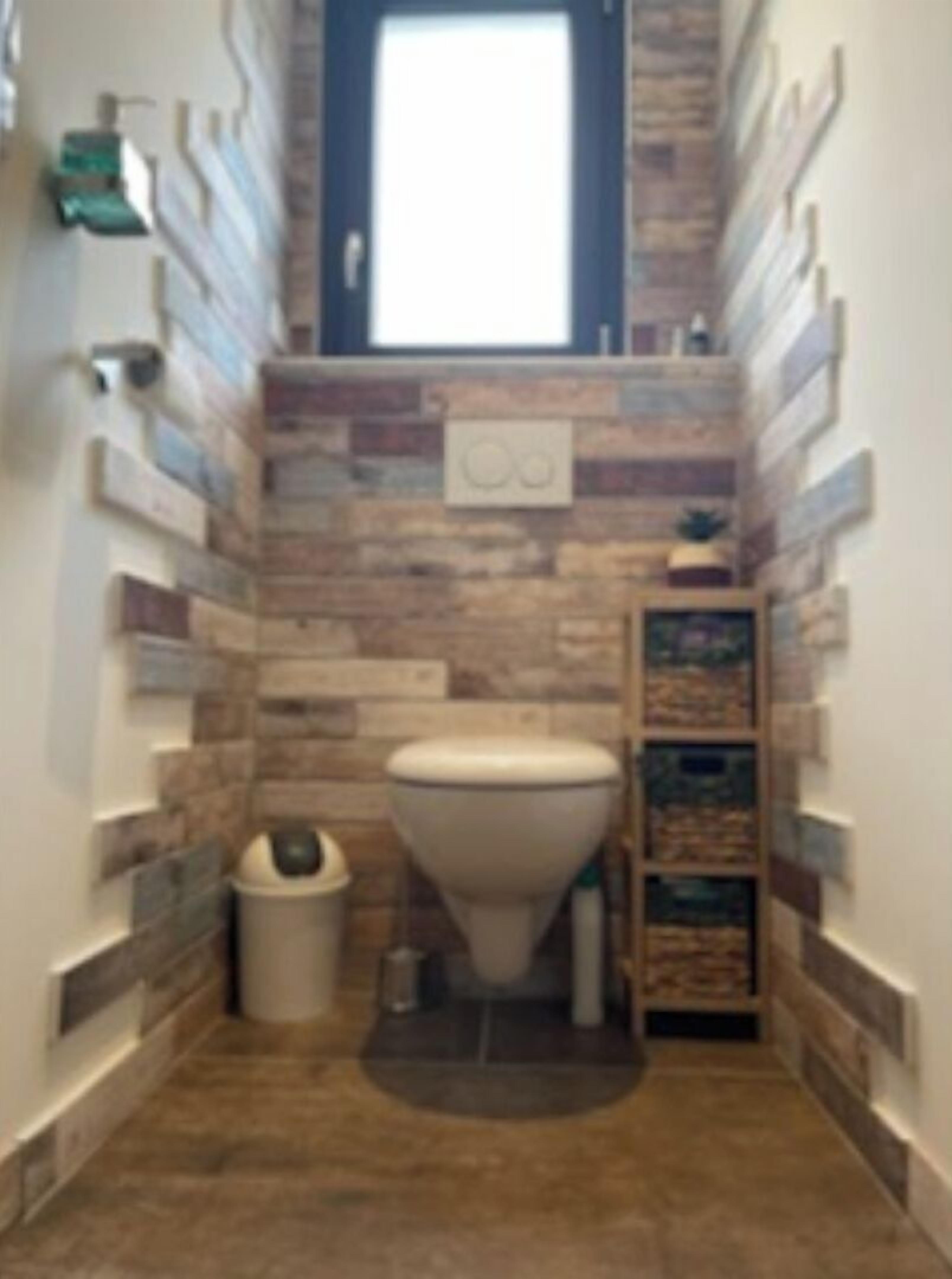 A narrow bathroom with a toilet, a trash bin, and a shelf with toiletries.
