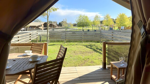 A wooden deck with a table and chairs, overlooking a fenced playground area with trees and a clear sky. - Camping Rural Montori (Ultramort)