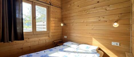 Une pièce de style cabane en bois, avec un lit, une fenêtre avec des rideaux et deux lampes fixées au mur.