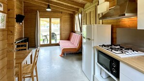 A wooden cabin interior with a kitchenette, a dining area with a table and chairs, and a sofa by the sliding glass door. - Camping Rural Montori (Ultramort)