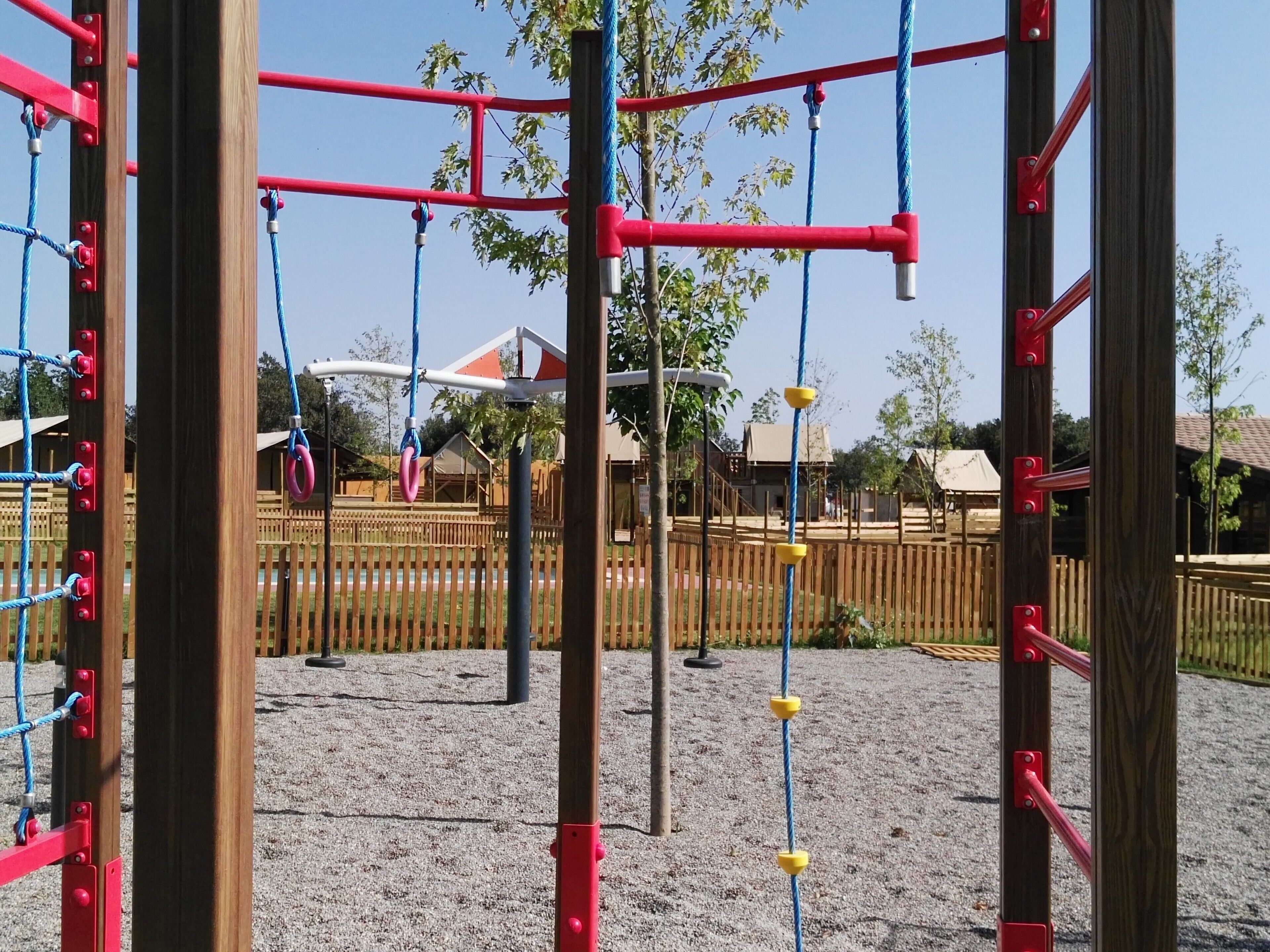 A playground with a wooden climbing structure, a fenced area, and a building in the background.