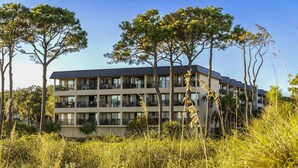 A multi-story hotel with balconies and a flat roof, surrounded by tall trees and grassy areas. - Oceanfront Hilton Head Island Condo w/ Pool! (Hilton Head Island)