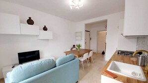 Una sala de estar moderna con un sofá azul, televisión, comedor con mesa y sillas de madera, y cocina con lavaplatos y estufa.