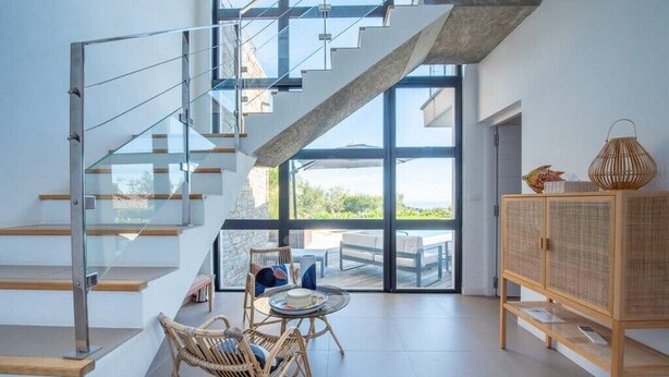A modern interior with a glass staircase, wicker furniture, and a view to the outdoors.