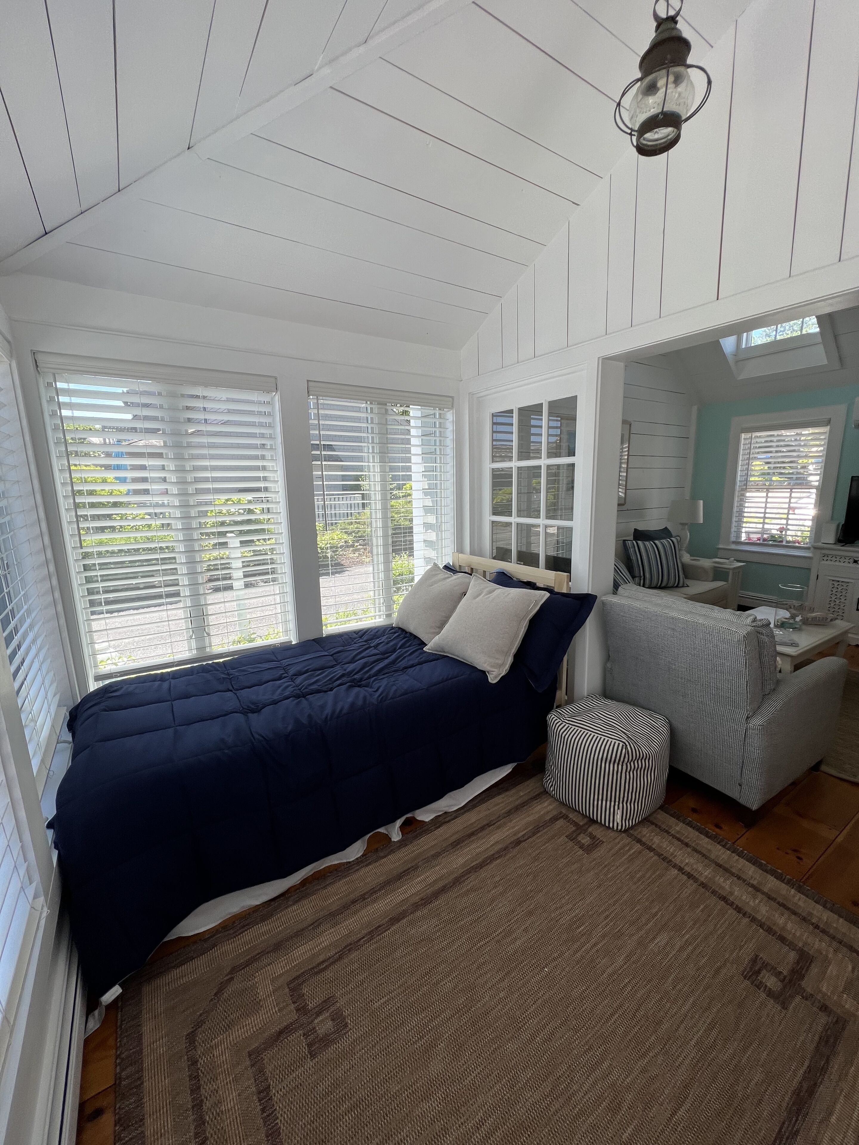 A bedroom with a large bed, a sofa, a small ottoman, and a window with blinds.