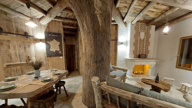 A rustic interior with a wooden dining table set for four, a fireplace, and a large tree trunk in the center. - Le Cul de Loup, self-catering cottage (Crouy-sur-Cosson)