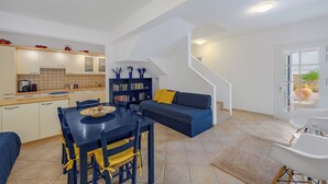Une cuisine moderne avec des meubles blancs, une table à manger avec des chaises, une bibliothèque et un canapé bleu dans un salon lumineux agrémenté d’un escalier.