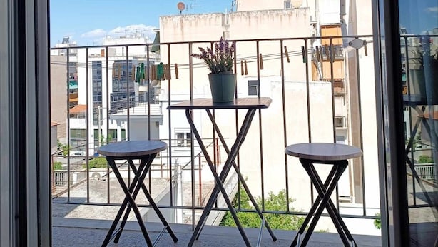 A balcony with two small tables and a potted plant, overlooking a cityscape with buildings and a clear sky. - Cozy Studio at Kallithea, Fully Equipped (Nea Smirni)