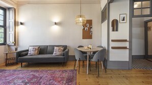 Ein modernes Wohnzimmer mit einem grauen Sofa, einem runden Esstisch mit Stühlen, einer hängenden Deckenleuchte und einem gemusterten Teppich.