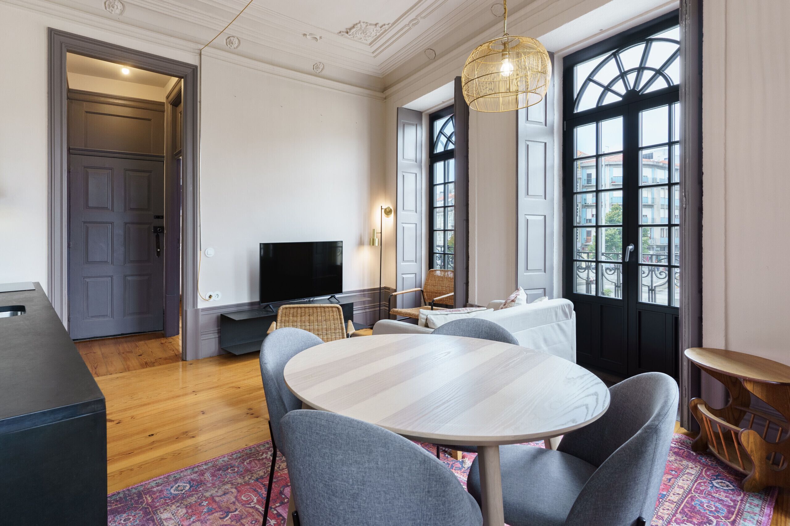 Een moderne woonkamer met een ronde tafel, grijze stoelen, een flatscreen-tv en een groot raam met uitzicht op gebouwen.