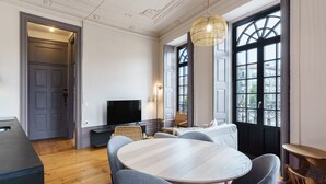 Una sala de estar moderna con una mesa redonda, sillas grises, un televisor de pantalla plana y un ventanal con vistas a edificios.