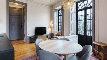 Een moderne woonkamer met een ronde tafel, grijze stoelen, een flatscreen-tv en een groot raam met uitzicht op gebouwen.