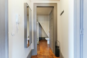 A narrow hallway with a wooden floor, a white wall-mounted phone, a black trash bin, and a door leading to a bathroom with a white toilet and a visible staircase.
