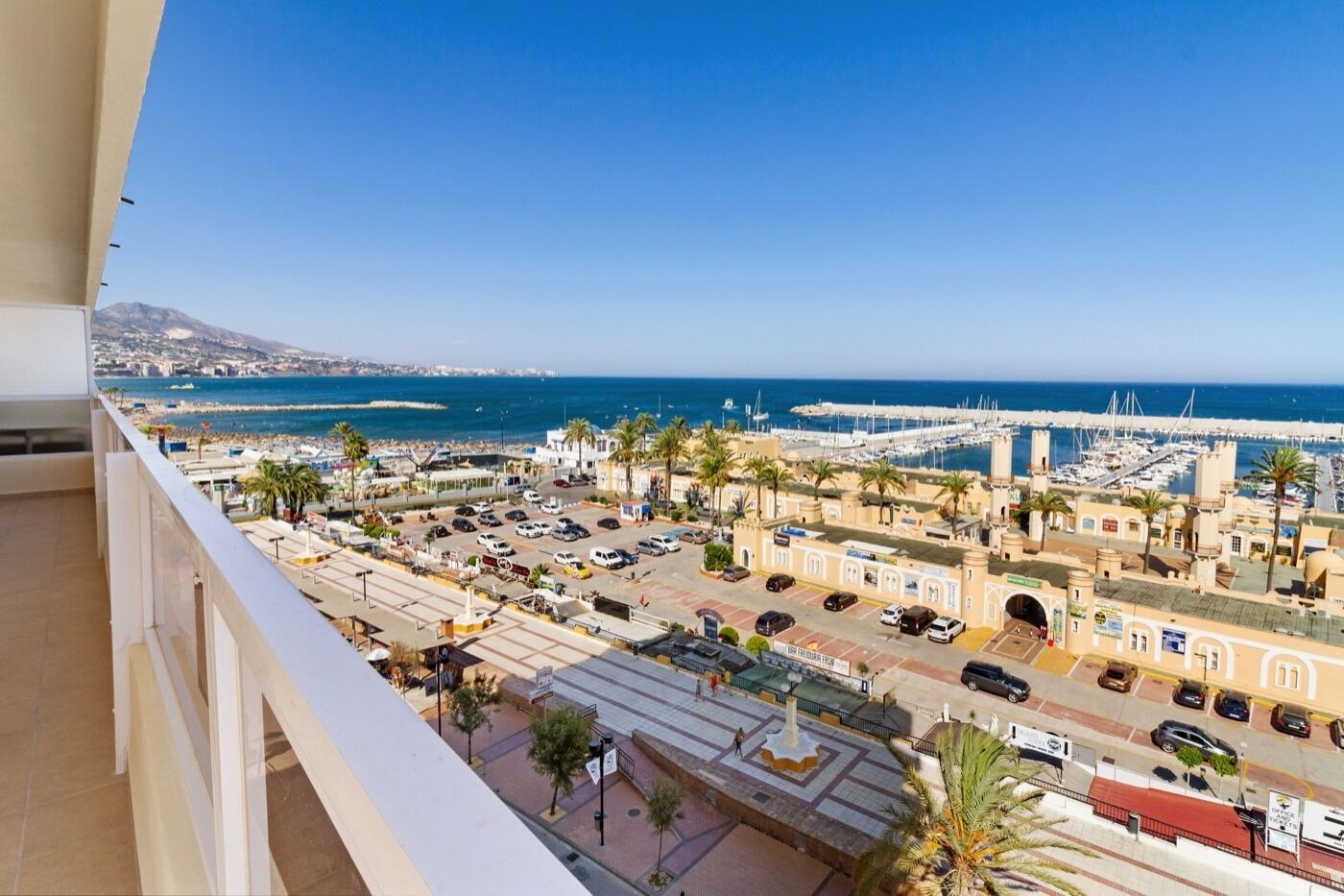 Een kuststadsgezicht met een jachthaven, geparkeerde auto's en gebouwen in mediterrane stijl.