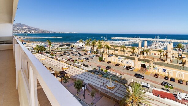 A coastal cityscape with a marina, parked cars, and buildings with Mediterranean architecture.