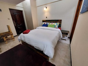 A bedroom with a bed, a wooden chair, a small table, and a wall-mounted light fixture.