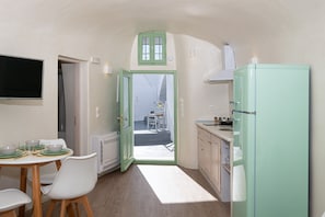 A compact kitchen with a dining area, a green-framed window, and a white refrigerator. - White Orchid Cave House & Studio (Santorini)