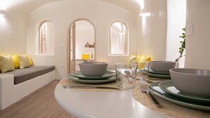 Un espace de salle à manger moderne avec une table blanche aux lignes courbes, des assiettes et des bols verts. On y trouve deux fenêtres en arc surmontées de coussins jaunes, posées sur un canapé blanc.