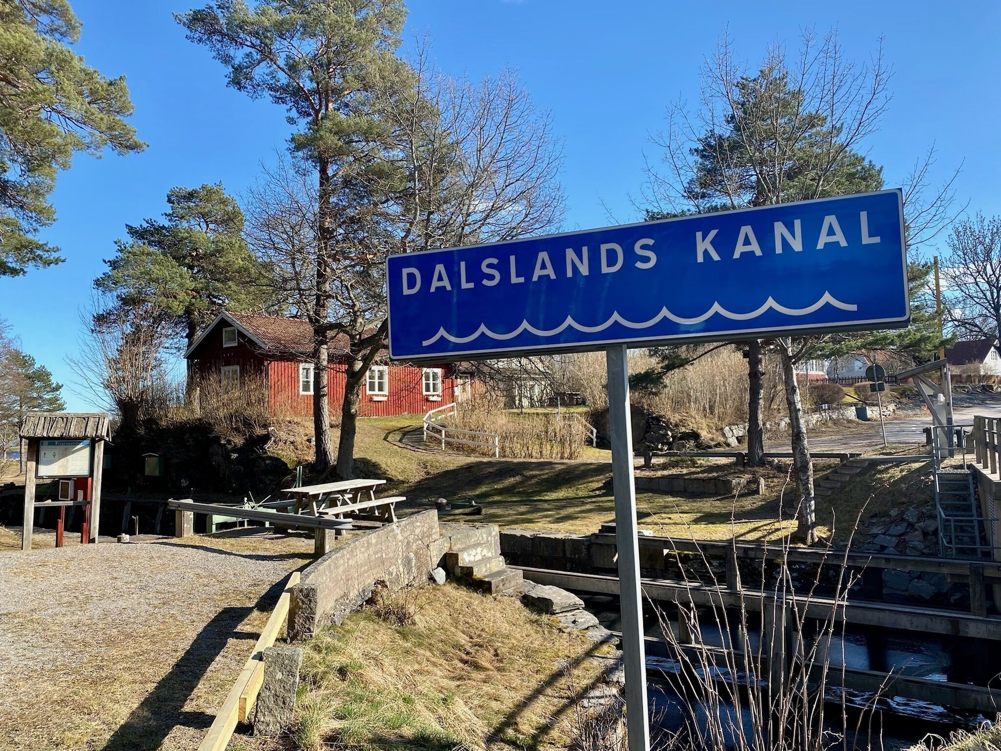 A sign for Dalslands Kanal with a wooden bench and picnic table nearby, a red house in the background, and a small dock.