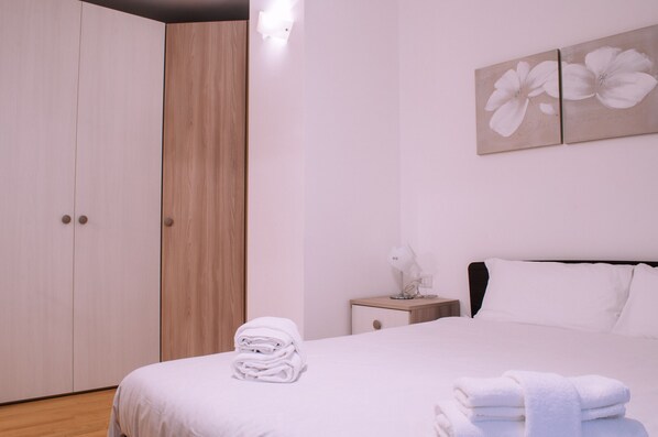 Una cama bien hecha con toallas blancas dobladas, una mesita de noche con una lámpara y una obra de arte en la pared con flores blancas.