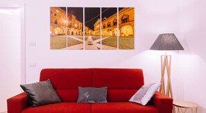 Un canapé rouge avec des coussins, un lampadaire et une œuvre d’art accrochée au mur représentant un bâtiment de nuit.