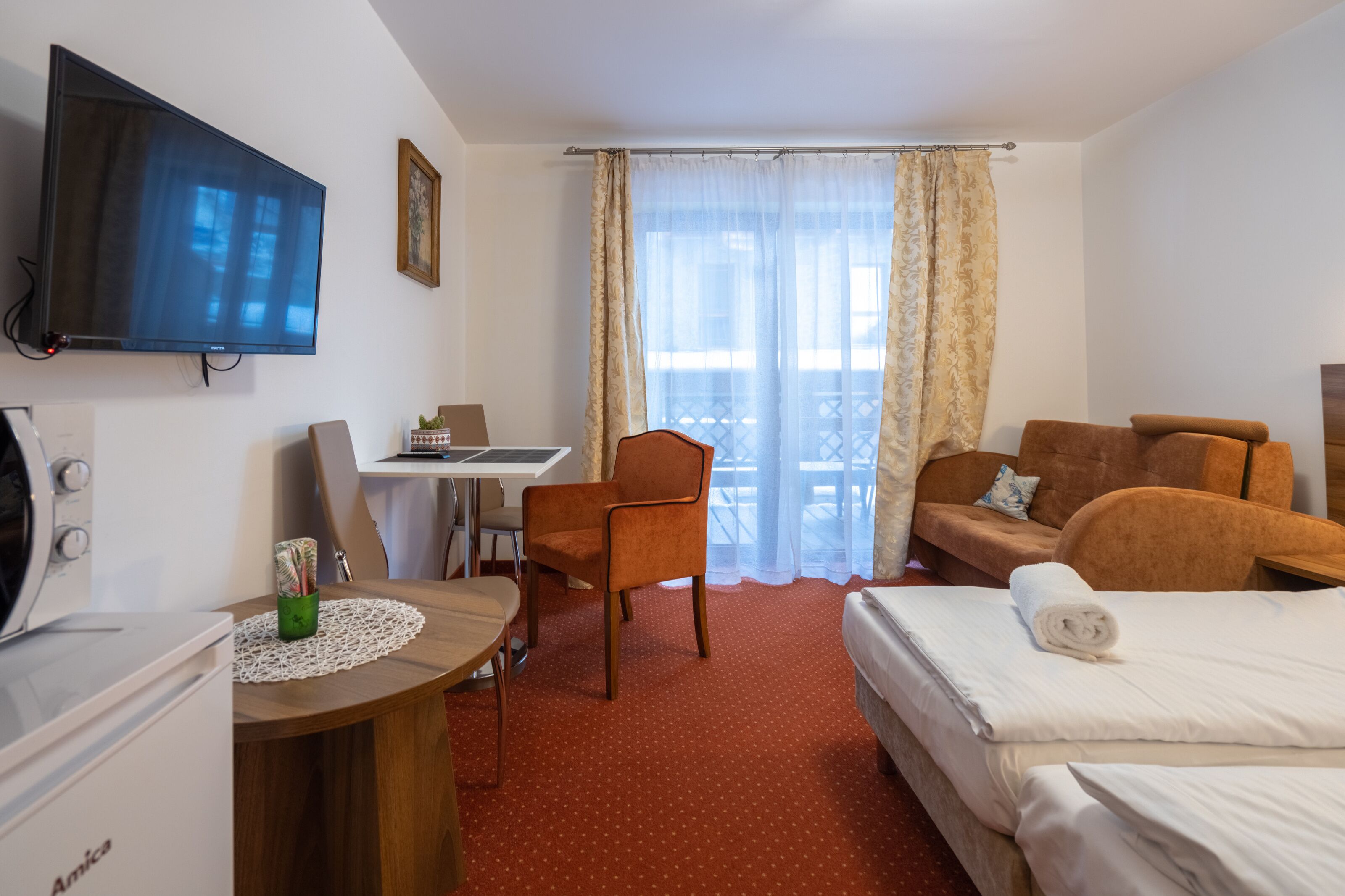 Habitación de hotel con cama, escritorio, silla, televisor y balcón con cortinas.