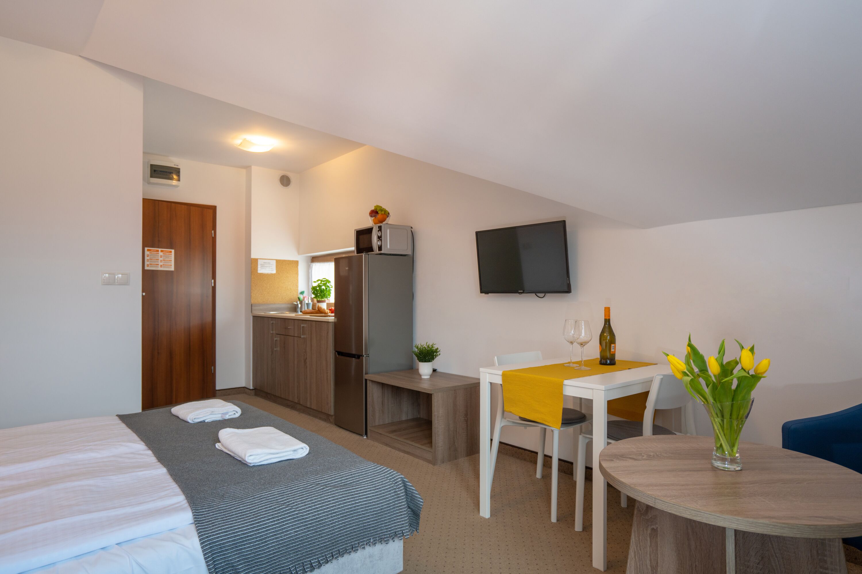Een compacte hotelkamer met een bed, een eettafel met stoelen, een kitchenette met een koelkast en magnetron, en een aan de muur bevestigde televisie.