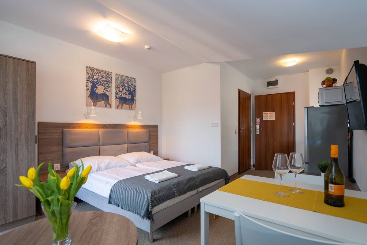 A hotel room with a bed, bedside lamps, a table with wine glasses, a bottle of wine, and artwork on the wall.