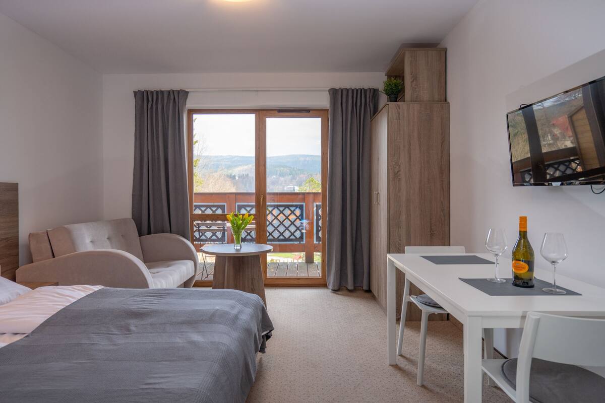 A hotel room with a bed, a sofa, a dining table with two glasses and a bottle, a TV on the wall, and a balcony with curtains.