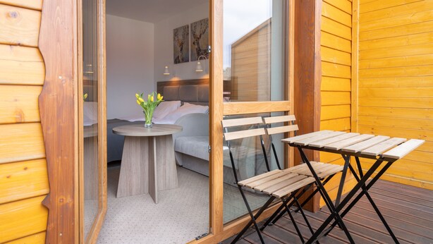A wooden deck with a table and chairs, a small round table with a vase of flowers, and a bed with a headboard in the background. - RentPlanet - Apartamenty Okrzei (Szklarska Poreba)