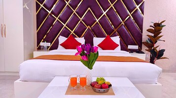 A hotel room with a large bed, a nightstand with a phone, a vase with flowers, two glasses of orange juice, and a fruit bowl.