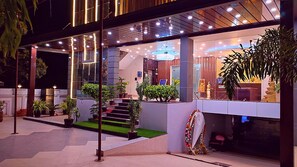 A modern hotel exterior with a glass facade, outdoor seating area, and a reception desk. - Hotel Aradhna Inn by ShriGo Hotels (Bakshi Ka Talab)