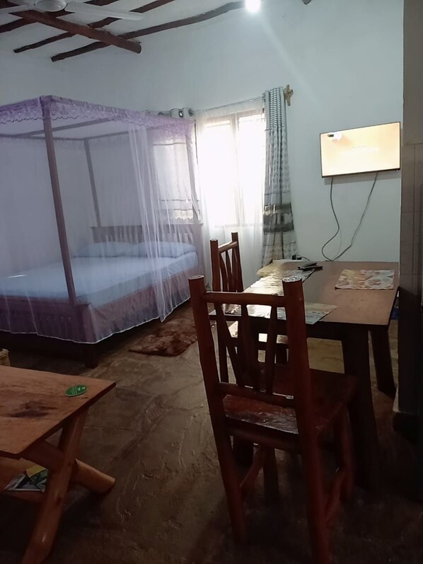 A room with a bed under a mosquito net, a wooden table with chairs, and a wall-mounted TV.