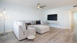 A modern living room with a sectional sofa, a glass coffee table, a ceiling fan, and a mounted TV.