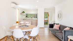A modern living room with a round wooden table, white chairs, a grey sofa, and a kitchen in the background. - Coastal Oasis - Pet Friendly - 6 Mins Walk to Beach (Callala Beach)