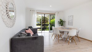 A modern living room with a grey sofa, a dining table with white chairs, and a large window with a view of greenery. - Coastal Oasis - Pet Friendly - 6 Mins Walk to Beach (Callala Beach)