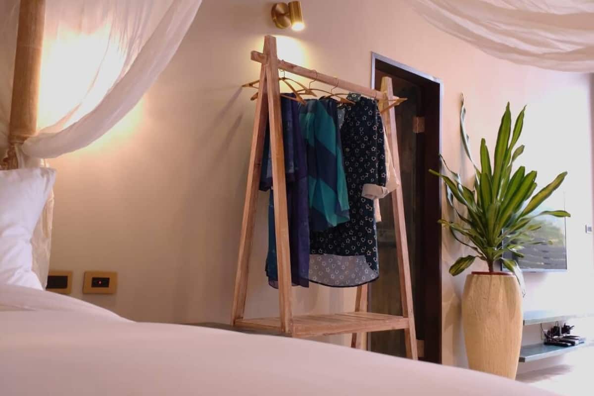 Ein Schlafzimmer mit Baldachinbett, einem Wäscheständer mit Kleidung, einem Topfblumenpflänzchen und einem an der Wand befestigten Fernseher.