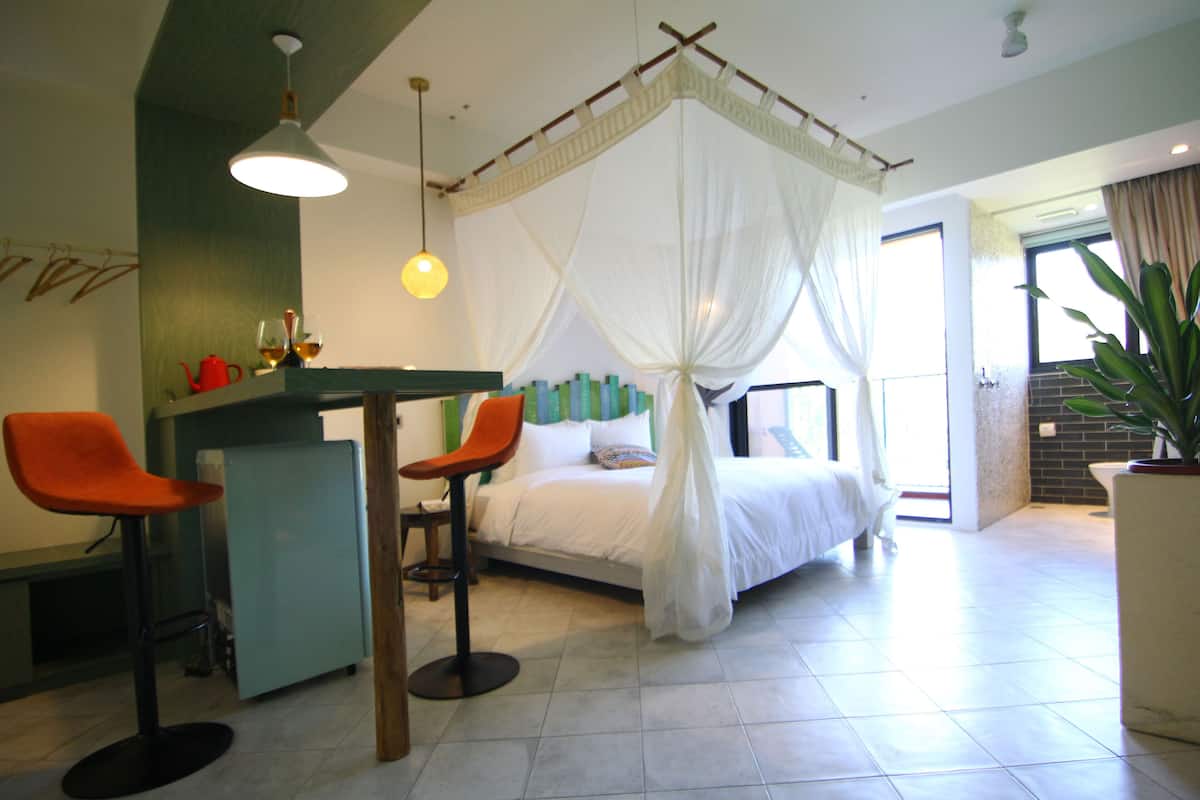 Ein Zimmer mit einem Bett, zwei Stühlen, einem Tisch, einem großen Fenster und einer Topfpflanze.